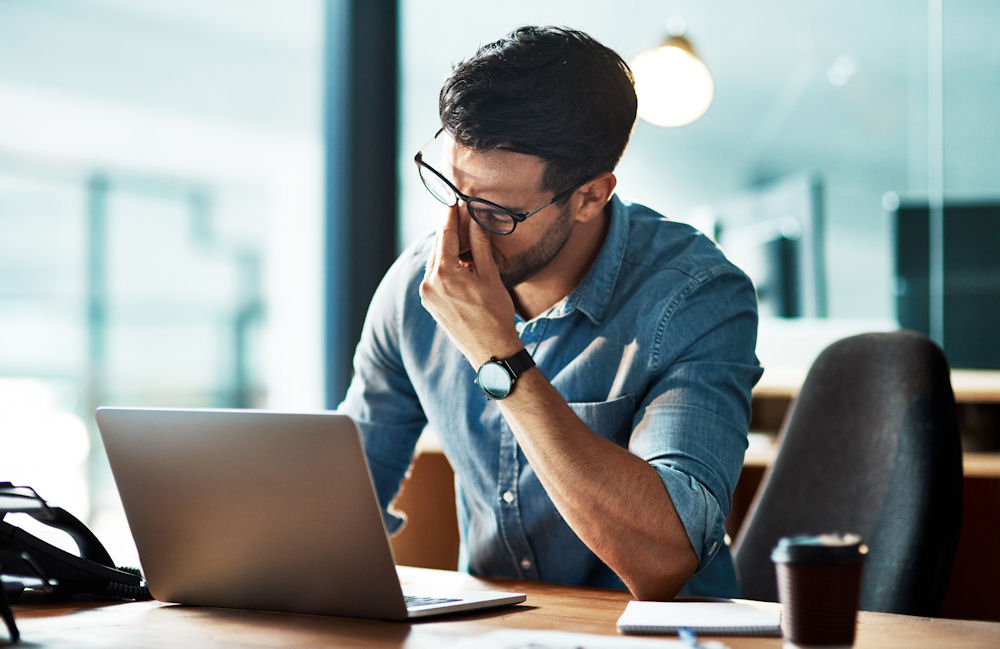 Man pinches bridge of nose in frustration while dealing with telehealth technology