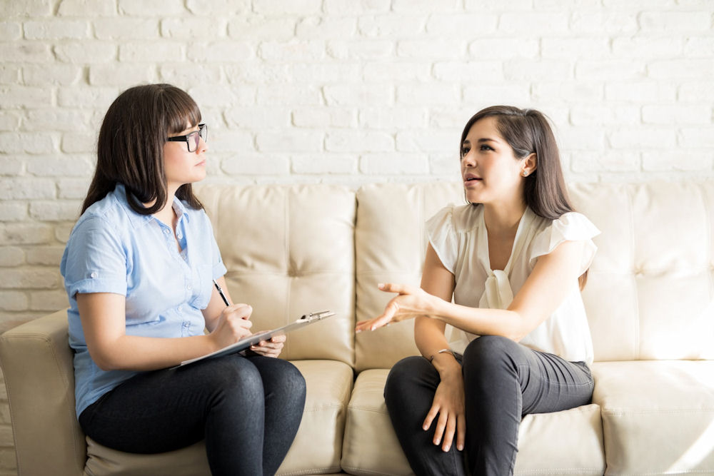 Woman describing thoughts on EMDR therapy while seated with therapist on white couch.