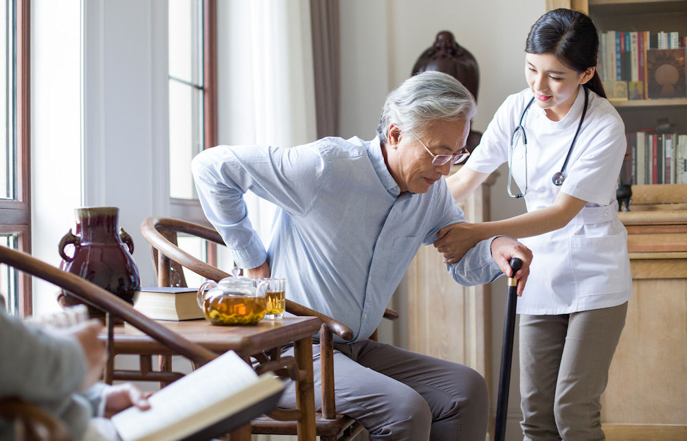 Home health care aide with stethoscope assists older man with can in rising up from wooden chair.
