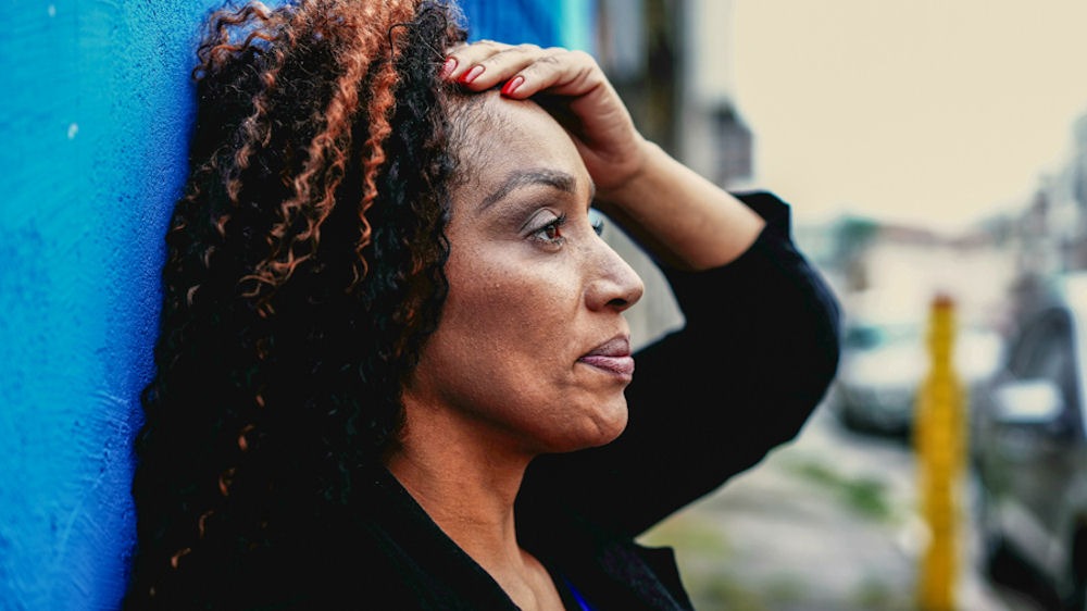 Middle-aged woman holds head and braces against wall in stress.