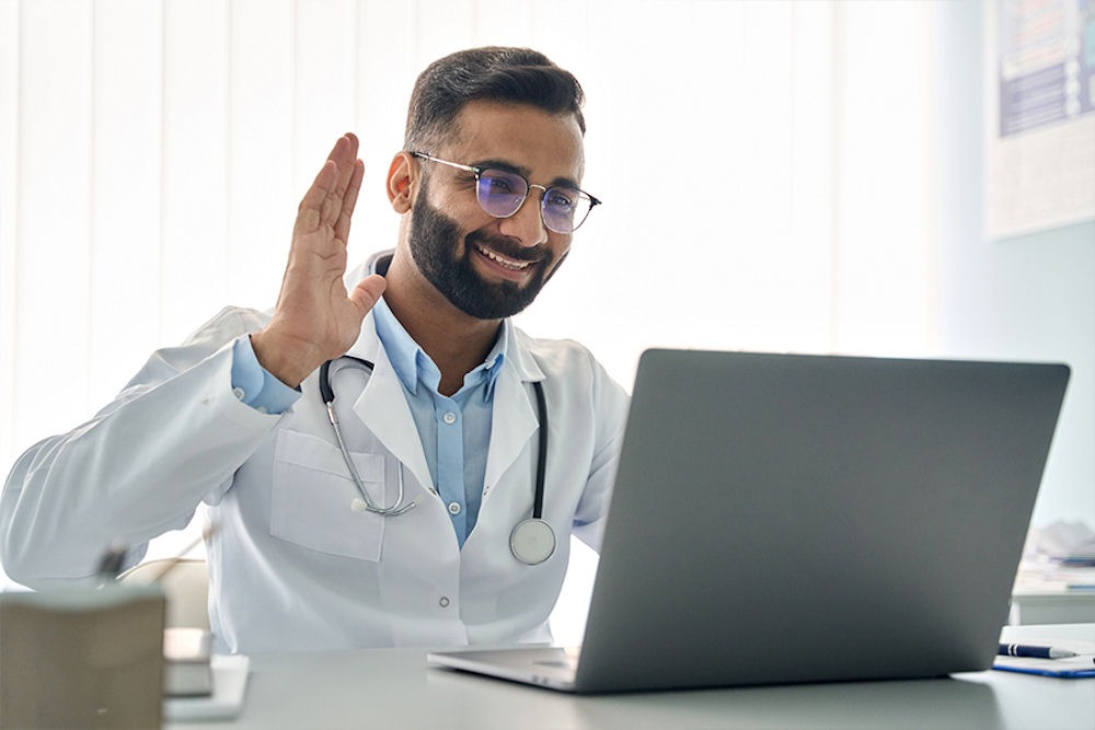 Doctor with glasses waves hello to patient over telehealth portal on laptop.