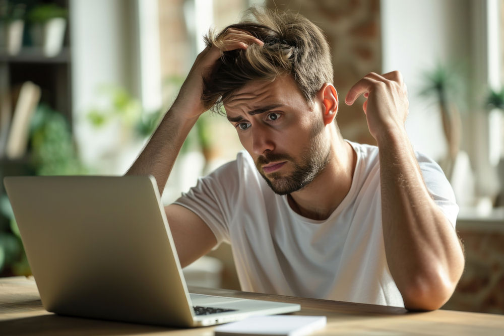 Confused man researches questions about anxiety on laptop with notepad.