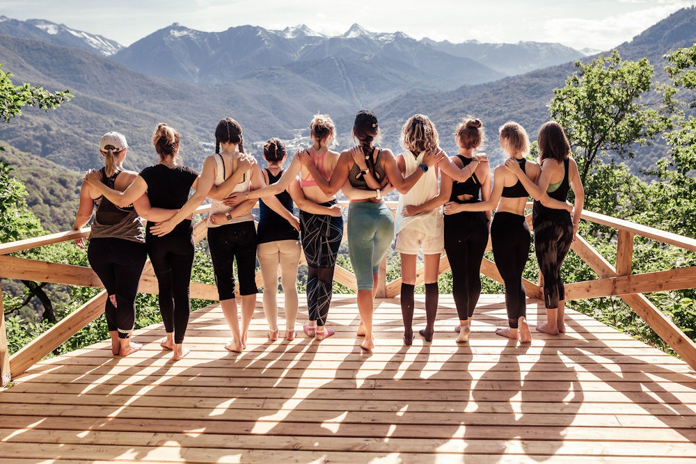 Rear view of ten women dress for yoga giving side hugs while looking out onto a valley from a balcony.