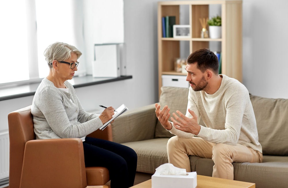 Man opening up to therapist with classes taking notes on notebook.