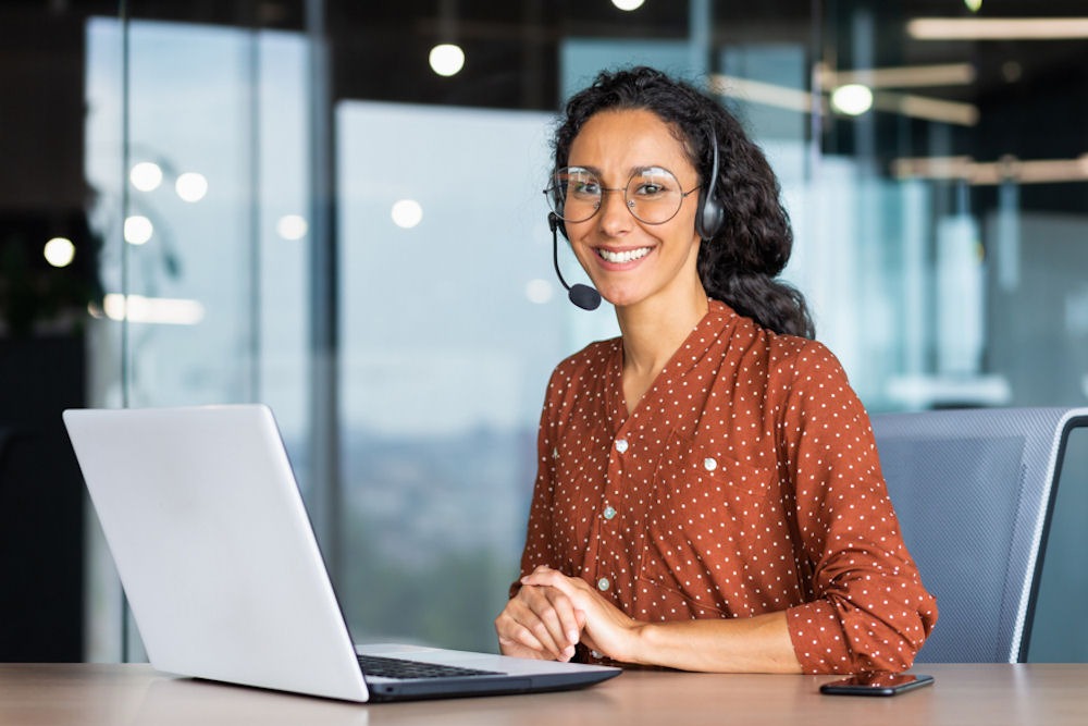 Lesbian woman smiling while working on laptop and wearing headset.